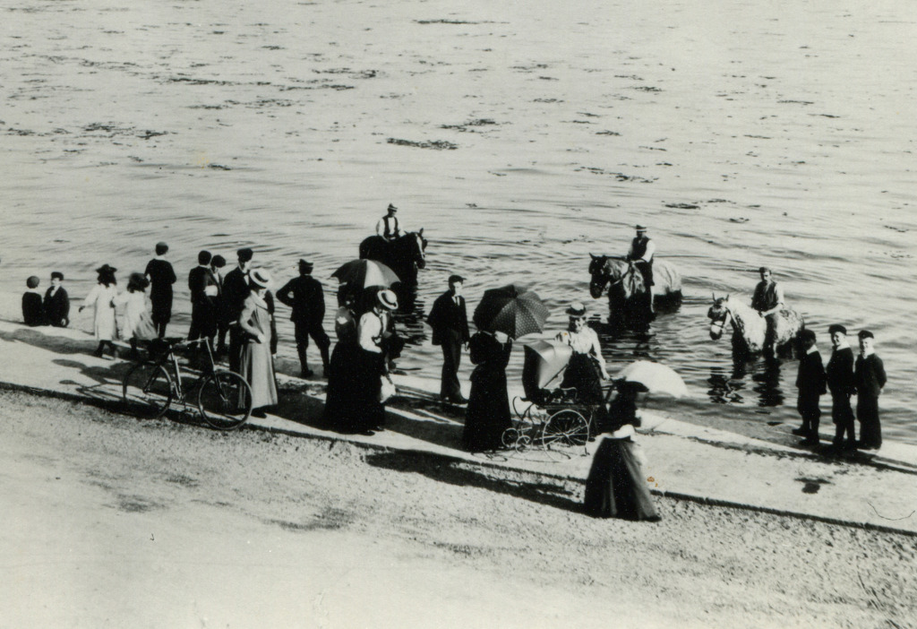 Detail: This image of 1910, shows people and horses enjoying the waters edge just outside the station. Views are across West Bay to the city walls and Town Quay. Image by kind permission of Southampton Daily Echo