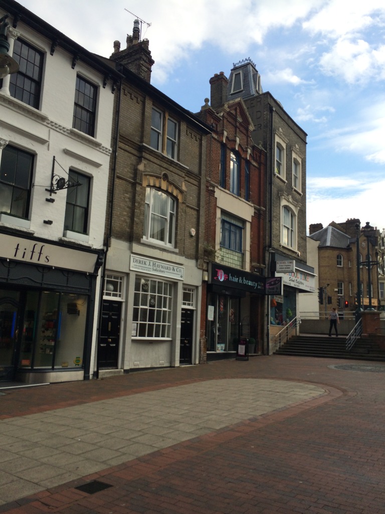 The retail units along the lower section of Railway Street, Chatham, appear busy and animated. The streetscape is abruptly interrupted and stifled by the A2 cutting and awkward steps, ramps and concrete wall. Image:Christopher Tipping